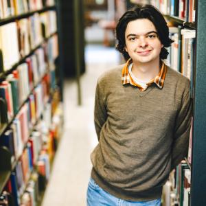 Jack Ciavolella stands in the stacks, leans against a bookshelf, and smiles at the camera.