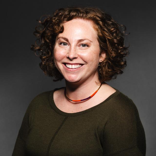 Farren Yero wearing a olive green shirt and an orange necklace smiling for her headshot with a black backdrop.