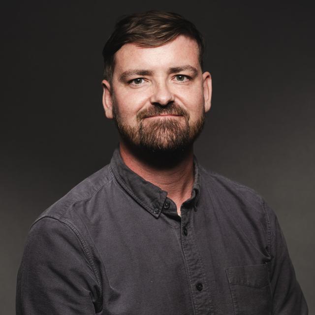 Owain Lawson wearing a grey button down shirt smiling for his headshot in front of a blackdrop.