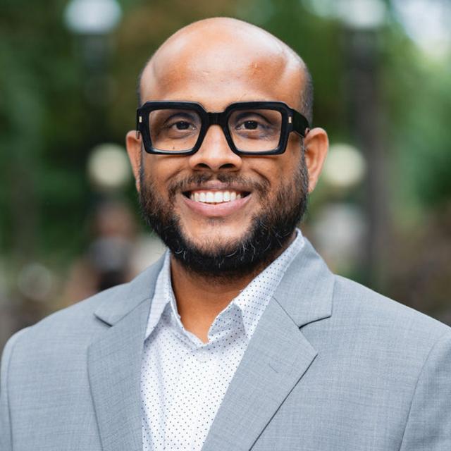 Sourav Ghosh wearing a grey suit jacket, white button-up shirt and glasses for his headshot outside.